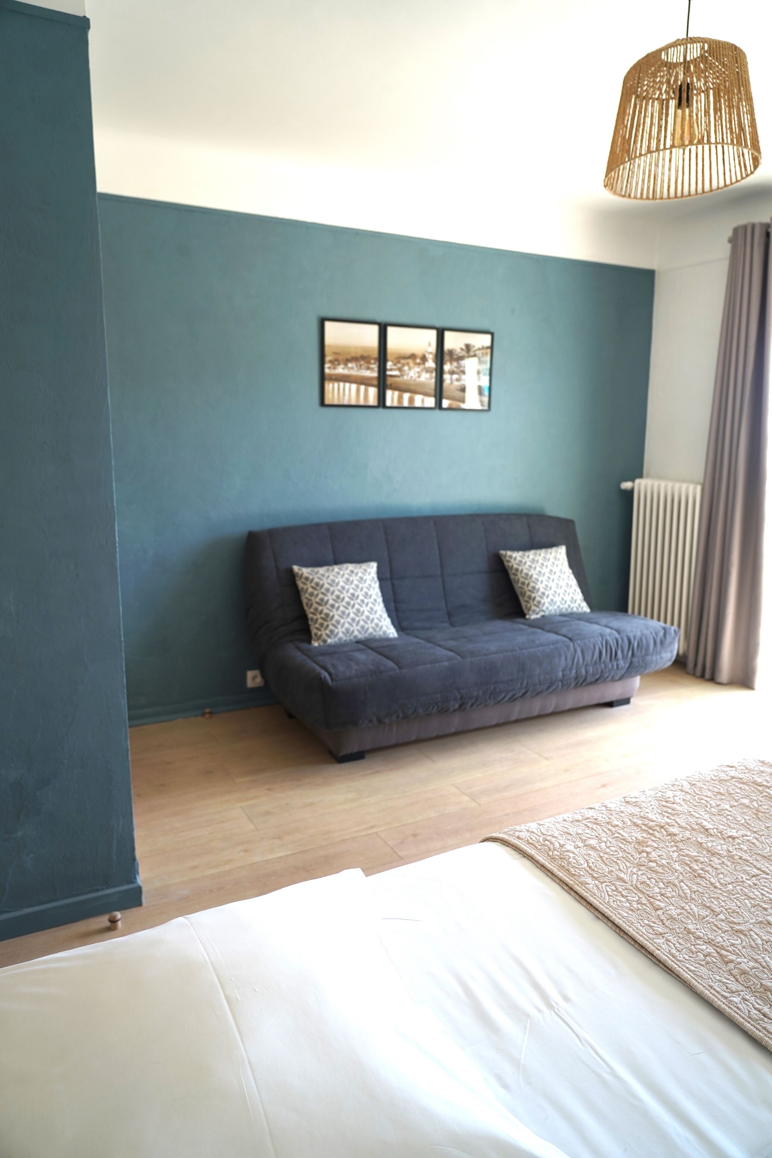 A room with a dark blue wall, a grey sofa with patterned cushions, a white bed, and a hanging woven light fixture.