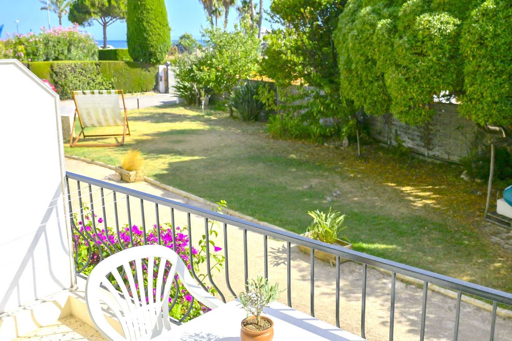 Un balcon avec une chaise blanche, un pot de fleurs et une vue sur un jardin bien entretenu, avec une pelouse, des arbustes et un mur de pierre.