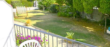 Un balcon avec une chaise blanche, un pot de fleurs et une vue sur un jardin bien entretenu, avec une pelouse, des arbustes et un mur de pierre.