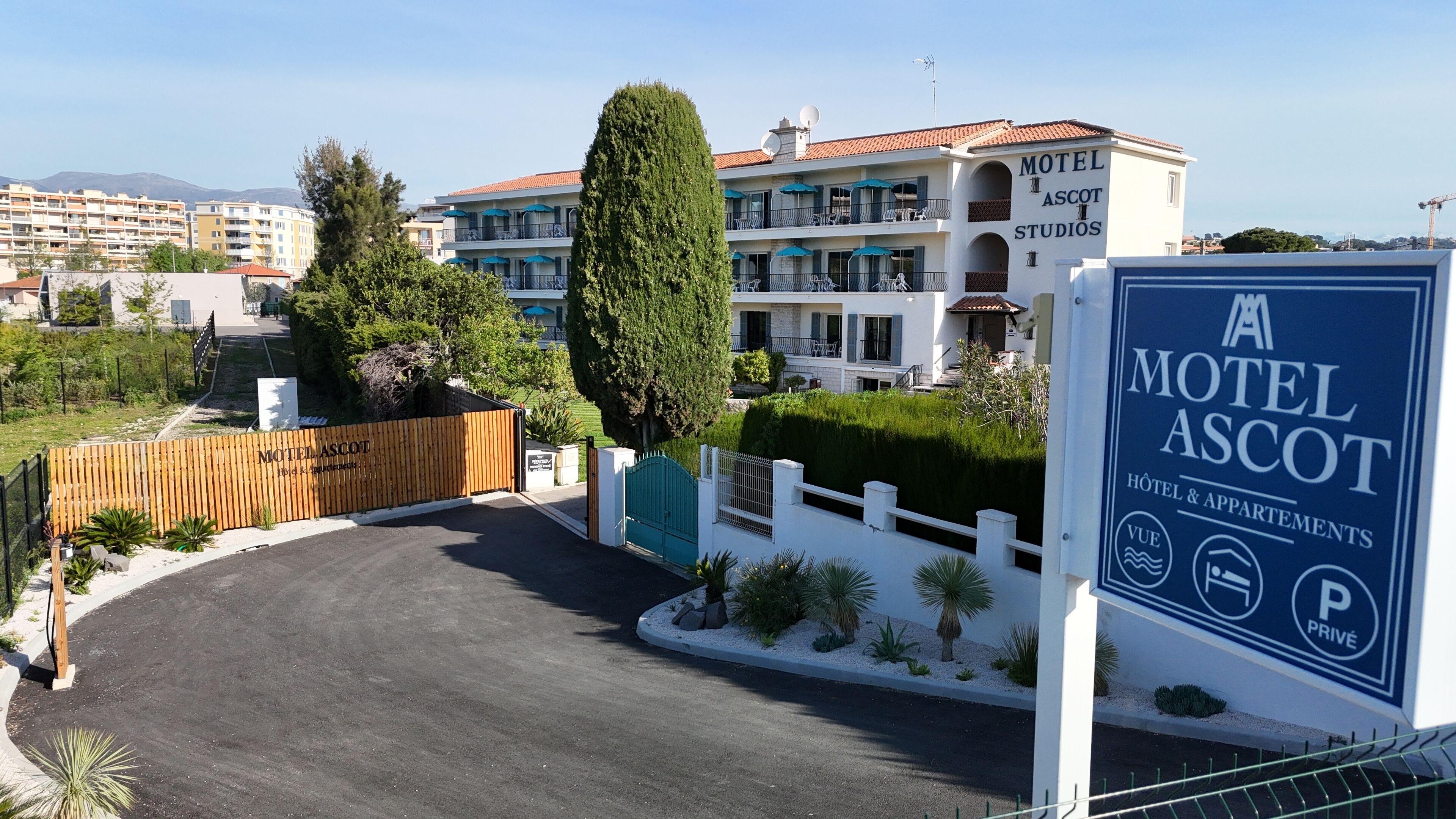 A hotel entrance with a gated driveway, a signboard for 'Motel Ascot', and a view of the surrounding buildings.