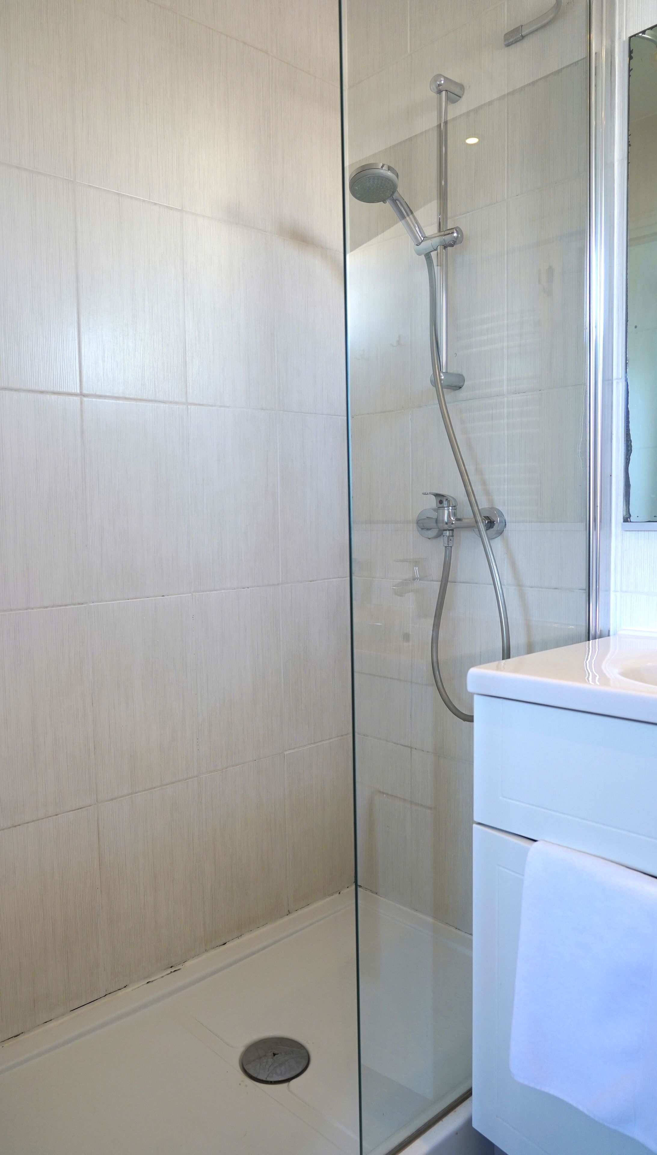 A glass-enclosed shower with a white tiled wall, a glass door, and a white sink with a towel.