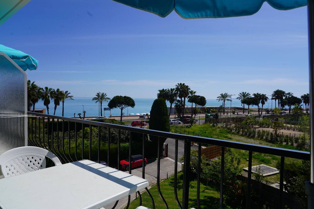 A balcony with a view of the sea, palm trees, and parked cars.