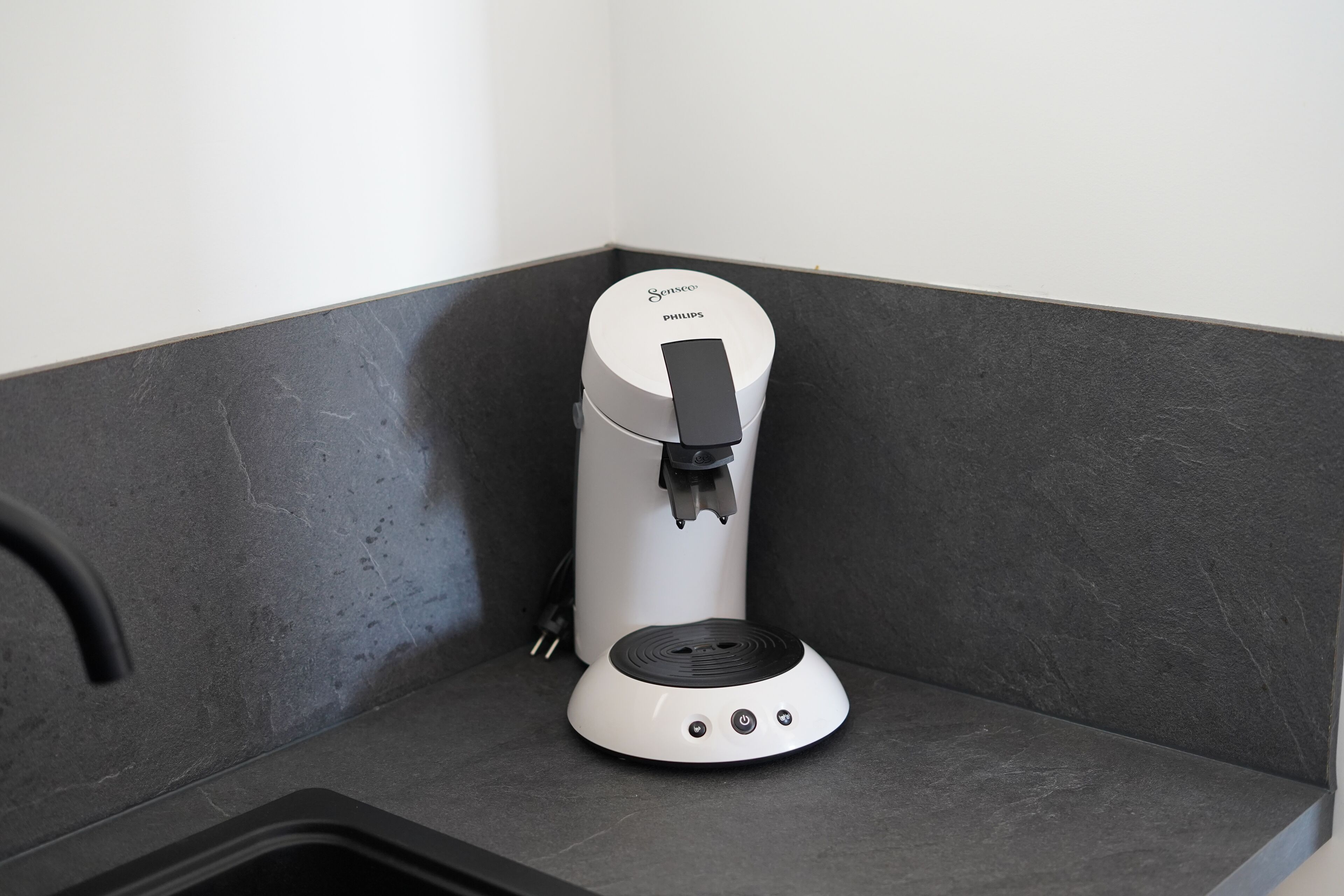 A coffee machine in a corner of a room with dark grey walls and a white baseboard.