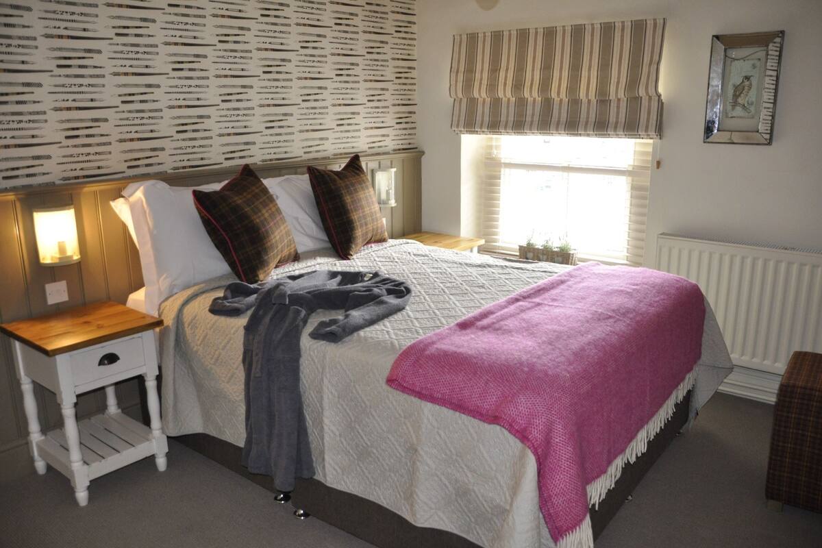 Quarto com cama, mesa de cabeceira e janela com cortinas.