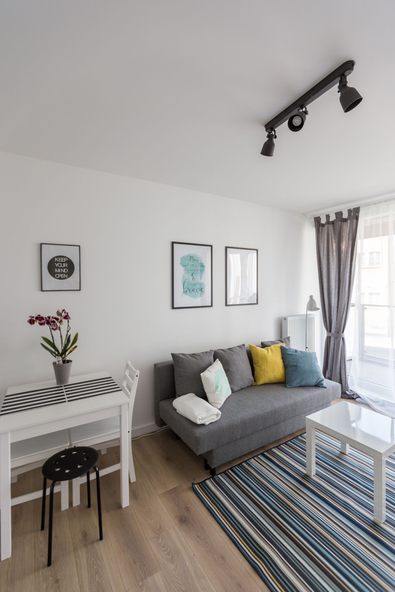 A modern living room with a grey sofa, a white coffee table, a striped rug, a white desk with a chair, and a potted plant.