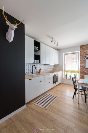 A modern kitchen with white cabinets, a black backsplash, and a wooden countertop. There is a window with blinds and a plant on the windowsill.