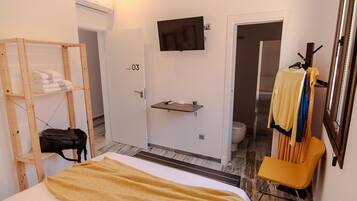 A hotel room with a bed, a wooden shelf with towels, a TV mounted on the wall, a small shelf with toiletries, and a chair.