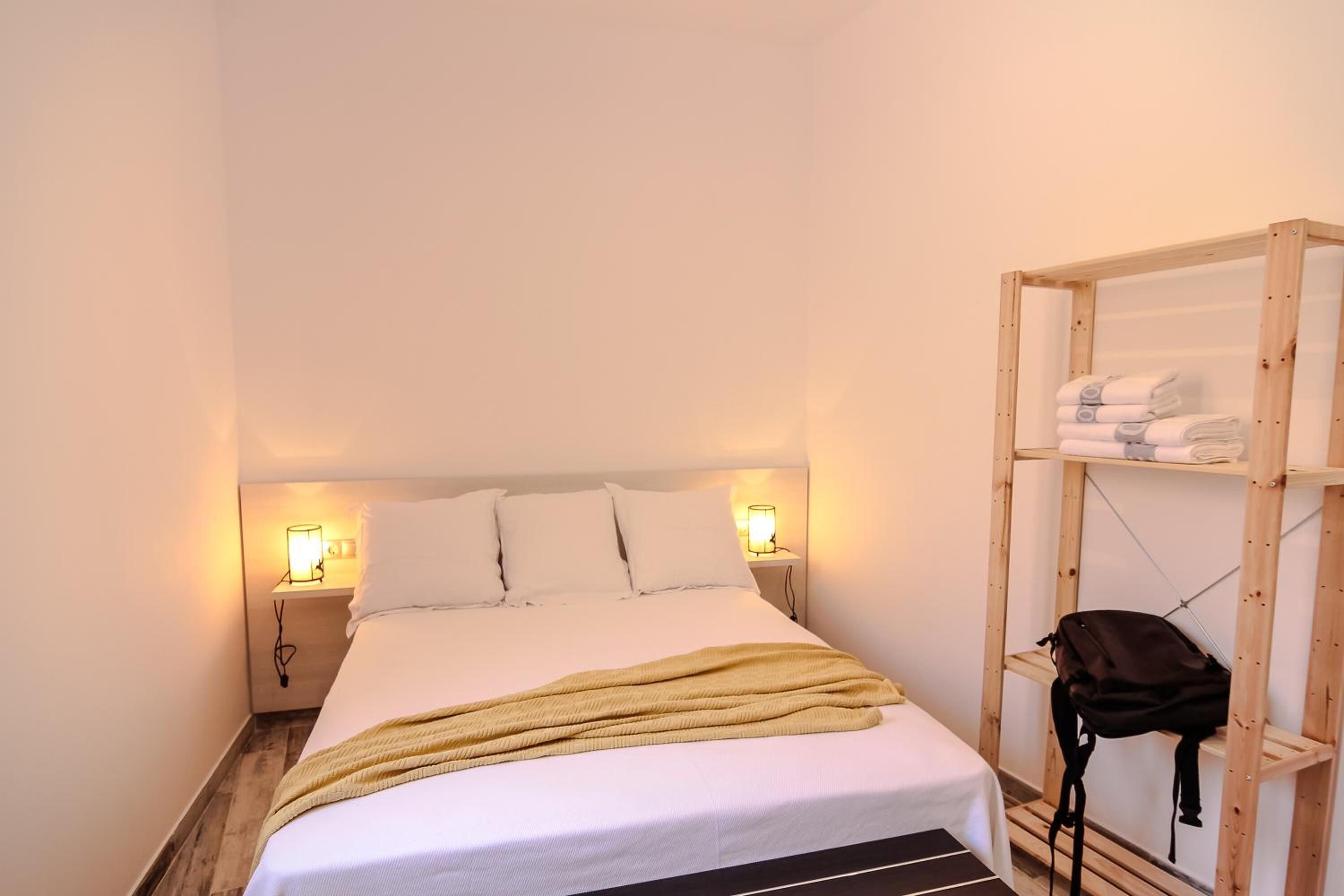 A neatly made bed with a white comforter and pillows, two bedside lamps, a wooden shelf with folded towels, and a backpack on the floor.