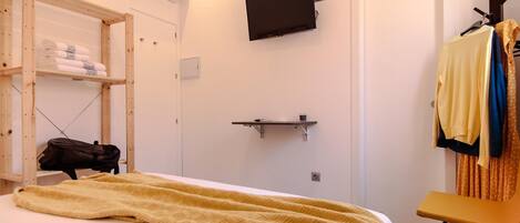 Une chambre avec un lit, un téléviseur fixé au mur, une étagère en bois avec des serviettes, une chaise et un porte-manteau avec des vêtements.