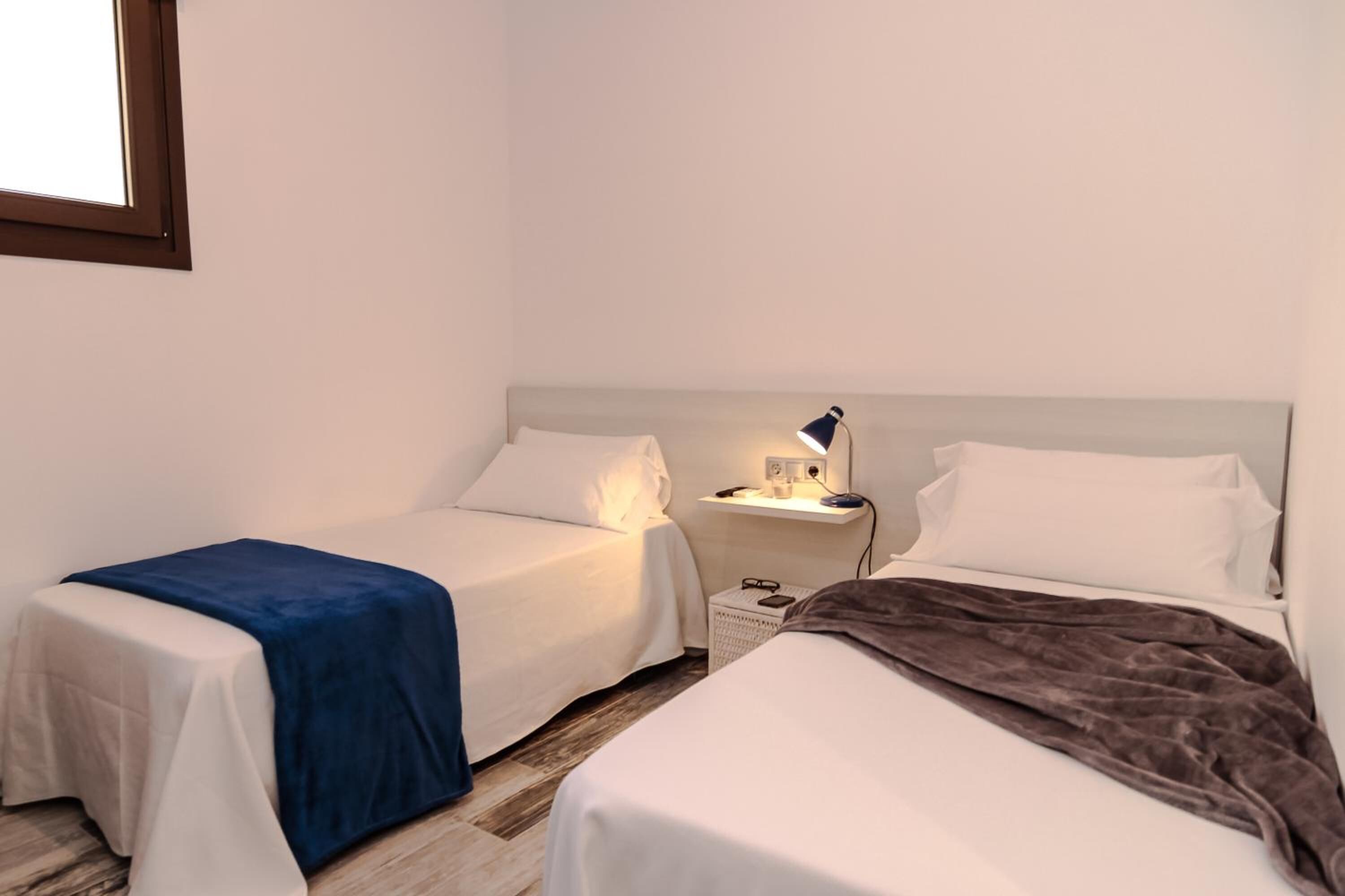 Un dormitorio con dos camas, una mesita de noche con lámpara y una ventana.