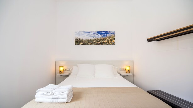Una cama bien tendida con sábanas blancas y una colcha color beige, dos mesitas de noche con lámparas, un cuadro enmarcado de un paisaje en la pared y un calefactor empotrado en la pared.