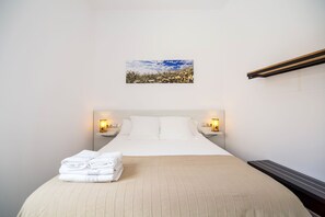 A neatly made bed with white linens and a beige bedspread, two bedside tables with lamps, a framed picture of a landscape on the wall, and a built-in heating element on the wall. - The 8 Rooms House (Tarifa)