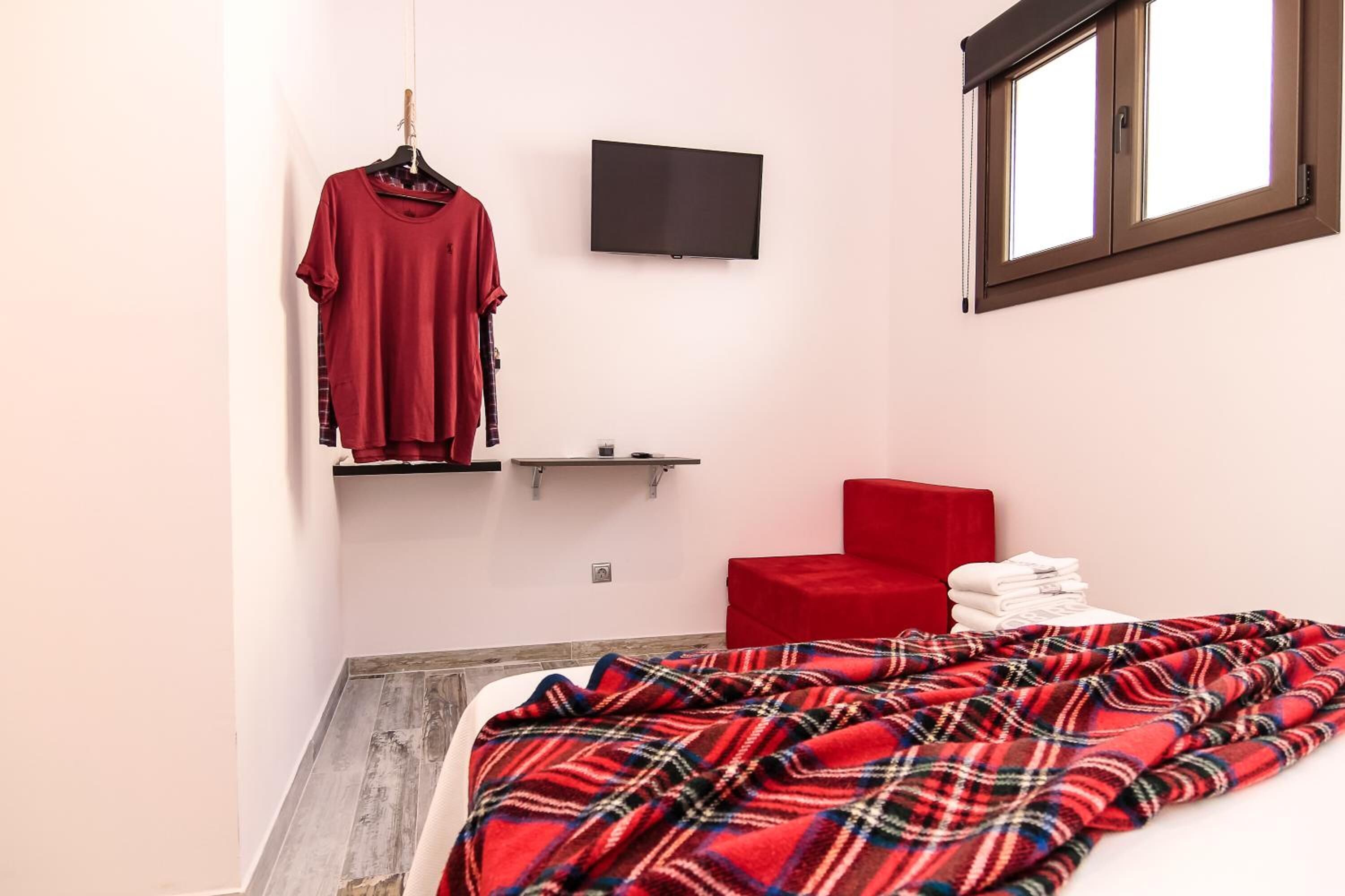 A bedroom with a bed, a red chair, a TV mounted on the wall, and a window with blinds.