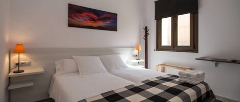 A hotel room with a bed, two bedside lamps, a painting, a window with a blind, and a small shelf.