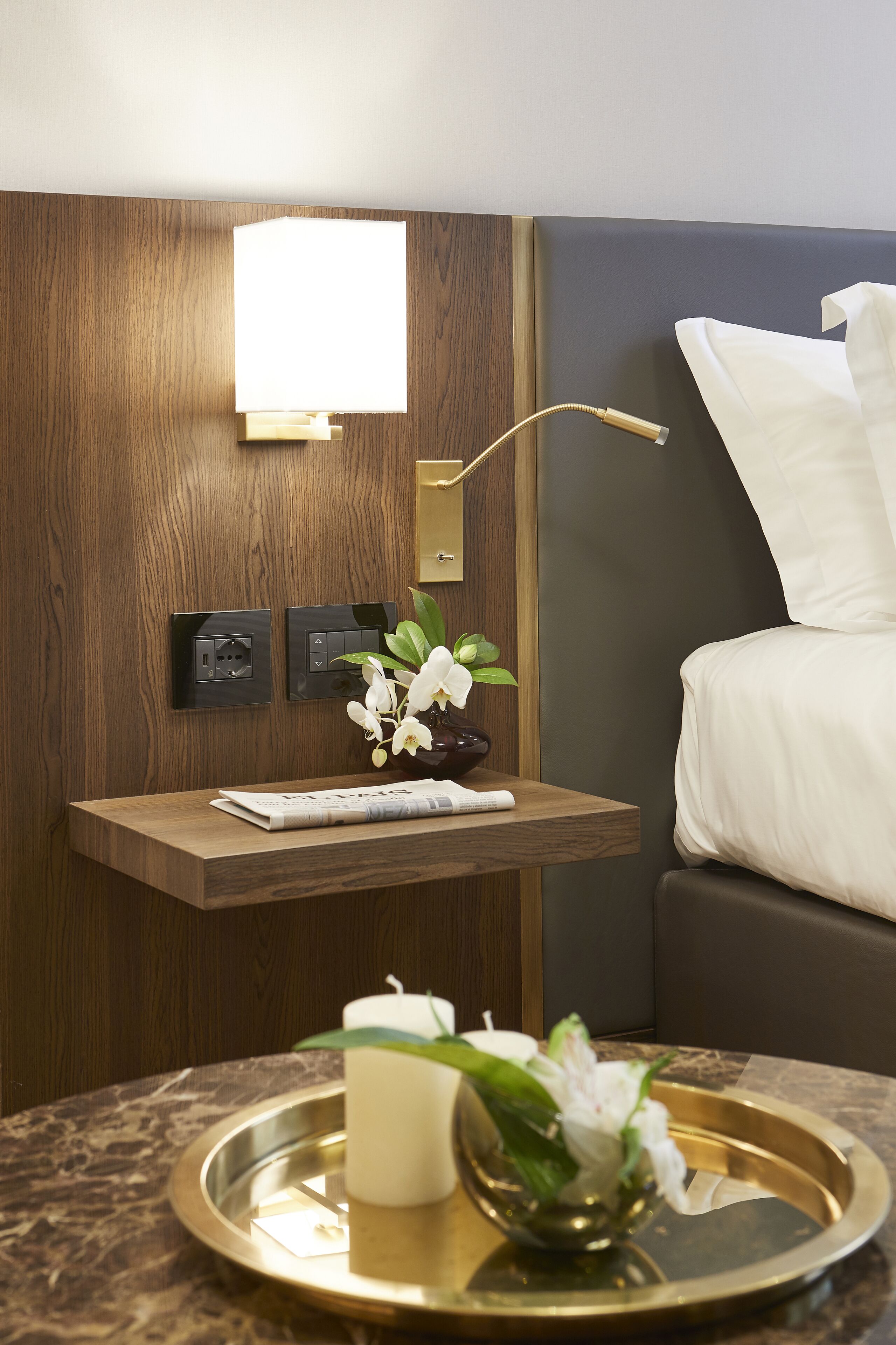 Habitación de hotel ordenada con una mesita de noche, una lámpara, una cama y una luminaria en la pared.
