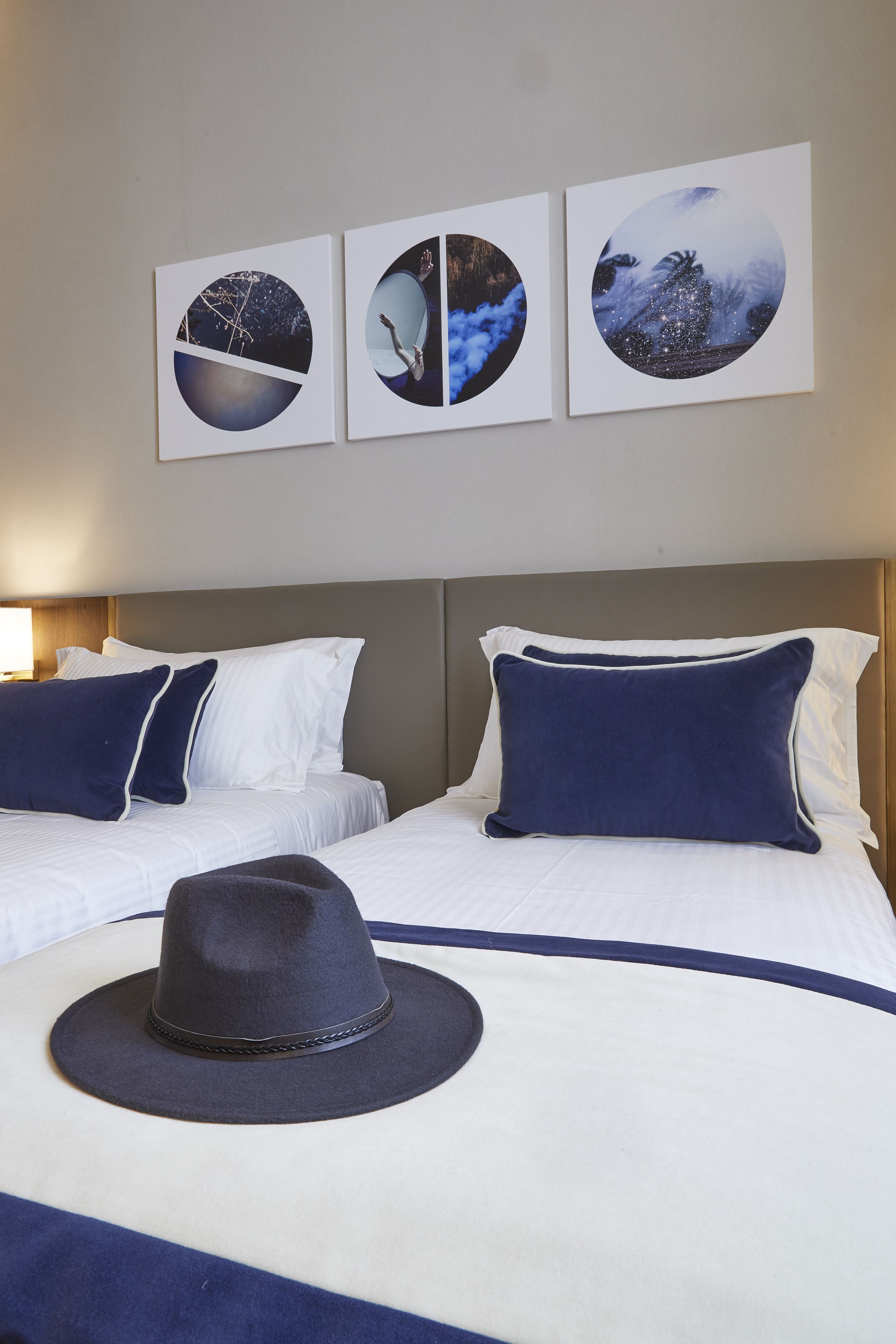 Une chambre d’hôtel avec deux lits, du linge de lit blanc et un chapeau bleu foncé posé sur l’un des lits. Trois œuvres d’art murales de forme circulaire sont accrochées au-dessus des lits.