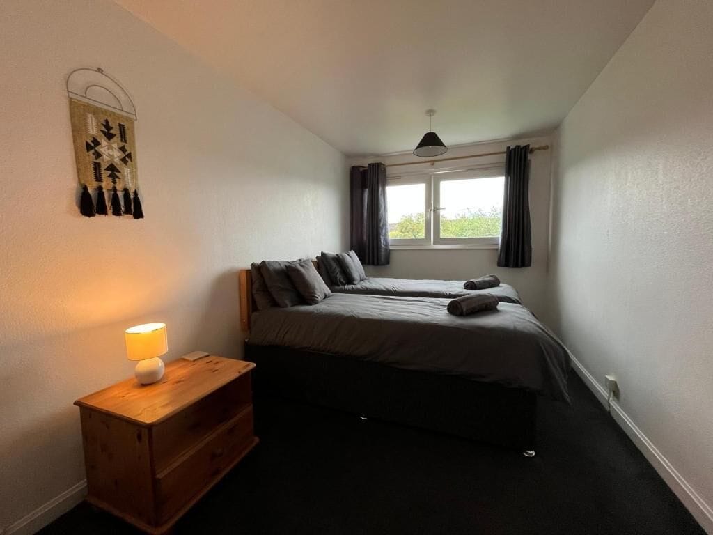 A bedroom with two beds, a bedside table with a lamp, a window with curtains, and a wall decoration.