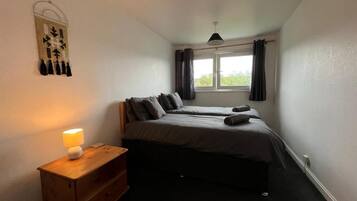 A bedroom with two beds, a bedside table with a lamp, a window with curtains, and a wall decoration.