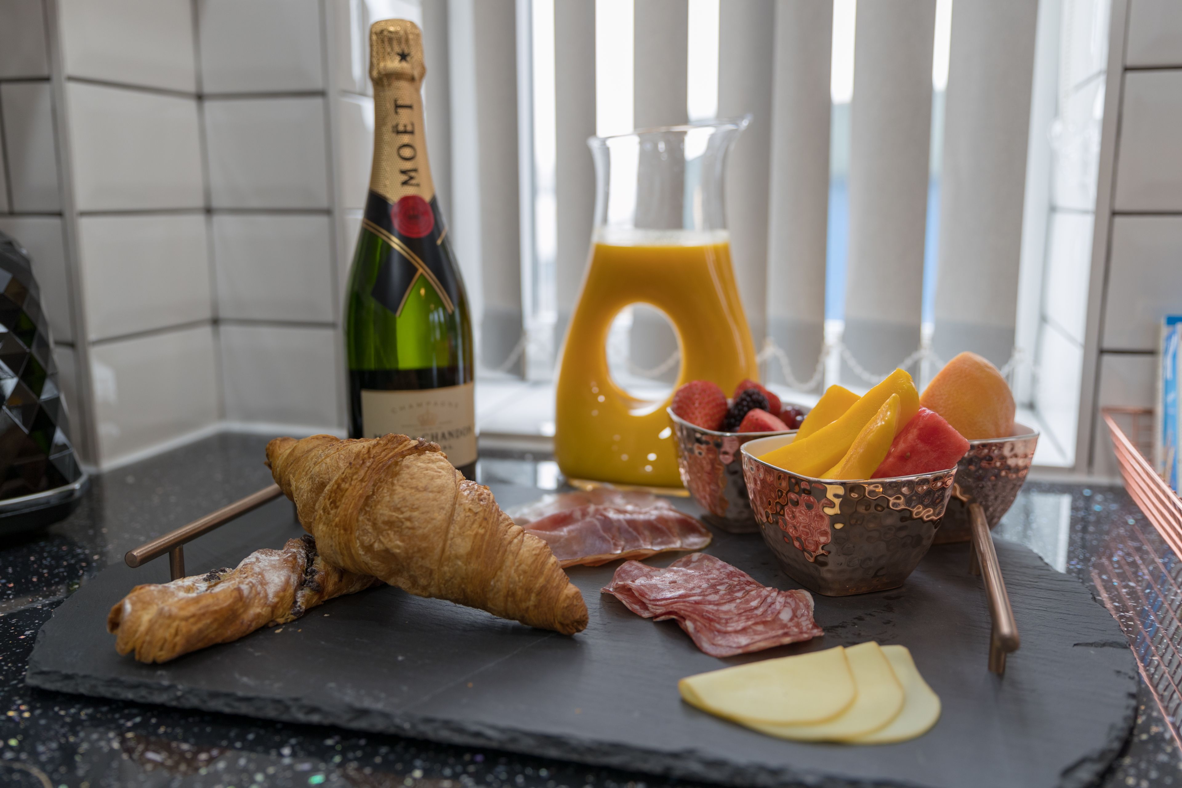A breakfast setup with croissants, cheese, fruits, and a bottle of champagne on a slate board.