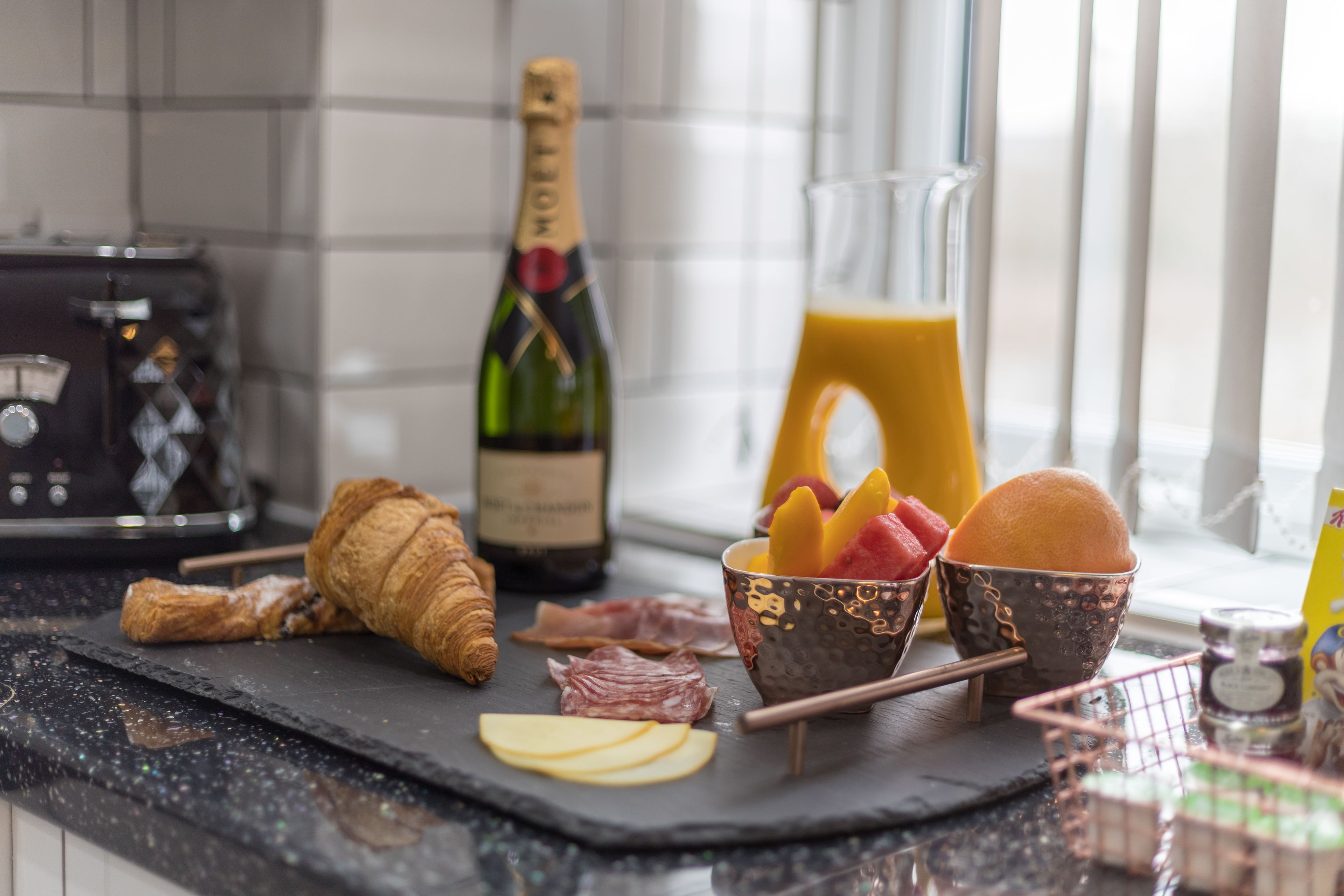 A kitchen counter with a croissant, cheese slices, fruit, and a bottle of champagne.