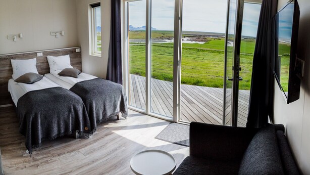 Un dormitorio con dos camas, un sofá y un amplio ventanal corredera que da a una terraza con vistas a un paisaje verde.