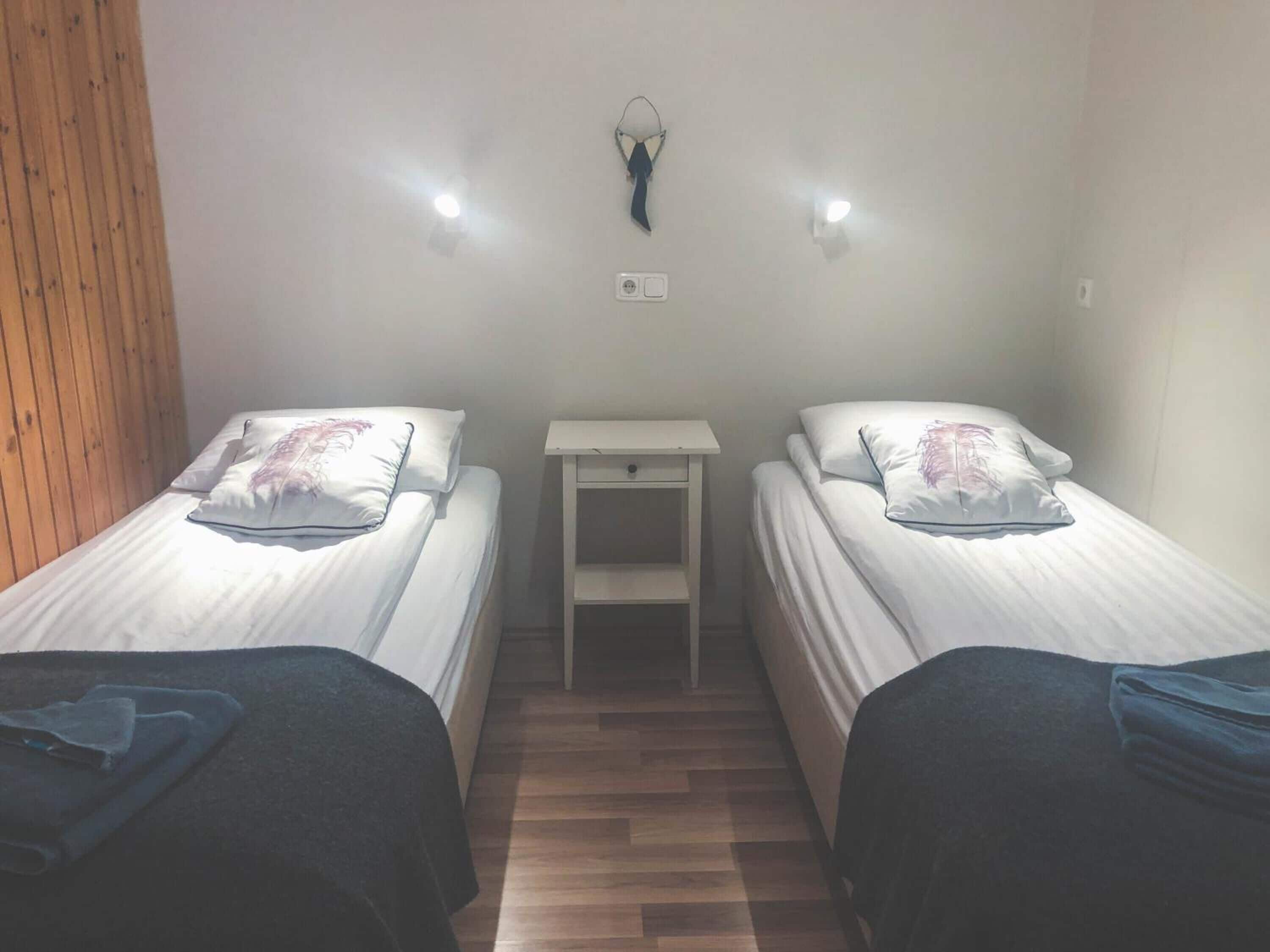 A room with two single beds, a small white nightstand, and a wall-mounted light fixture.