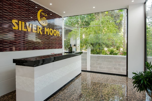 Un espace d’accueil au design moderne, comprenant un comptoir de réception, une grande fenêtre donnant sur un paysage verdoyant et un mur orné de l’inscription « Villa Silver Moon ».