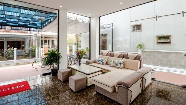 A modern hotel lobby with a sofa, ottoman, and coffee table. There is a red welcome mat, a potted plant, and a bicycle outside. - Silver Moon Villa Hoi An (Da Nang)