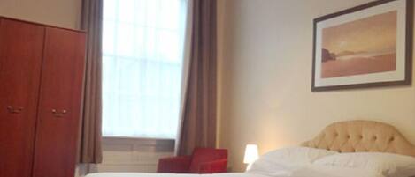 A bedroom with a bed, a wardrobe, a window with curtains, a wall-mounted picture, and a bedside lamp.