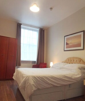 Un dormitorio con cama, armario, ventana con cortinas, un cuadro en la pared y una lámpara de noche.