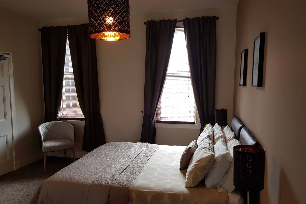 A bedroom with a bed, a chair, a bedside table, a lamp, and two windows with curtains.