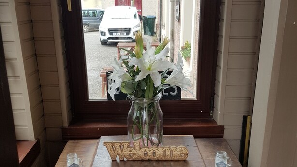Un escritorio de madera con un cartel de 'Welcome', un florero con flores blancas y una taza.