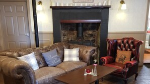 Una habitación acogedora con un sofá de cuero, una mesa de centro de madera y un sillón rojo. Se observa una chimenea con detalles de piedra al fondo.