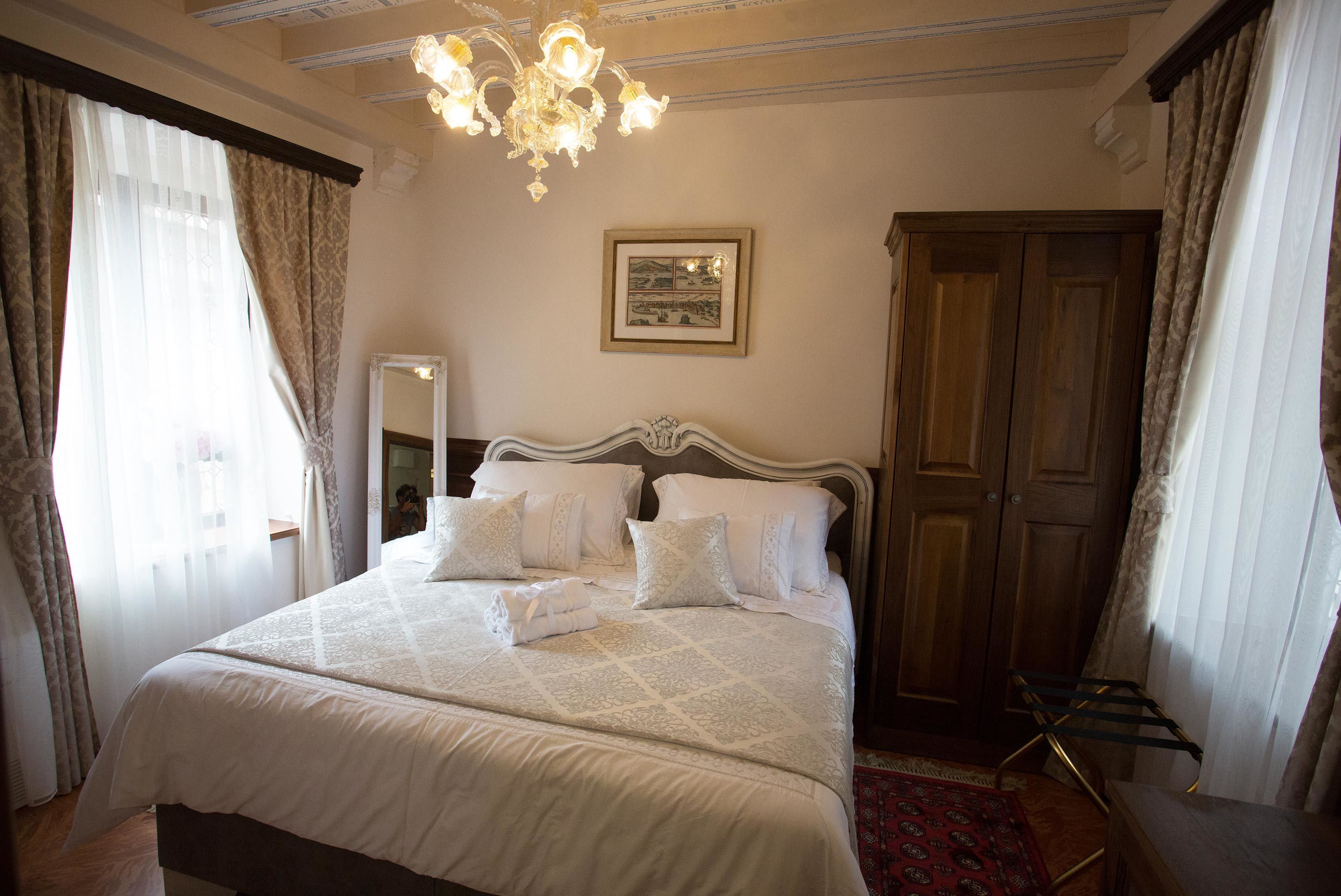 A neatly made bed with white linens, a chandelier, a wooden wardrobe, and a mirror on the wall.