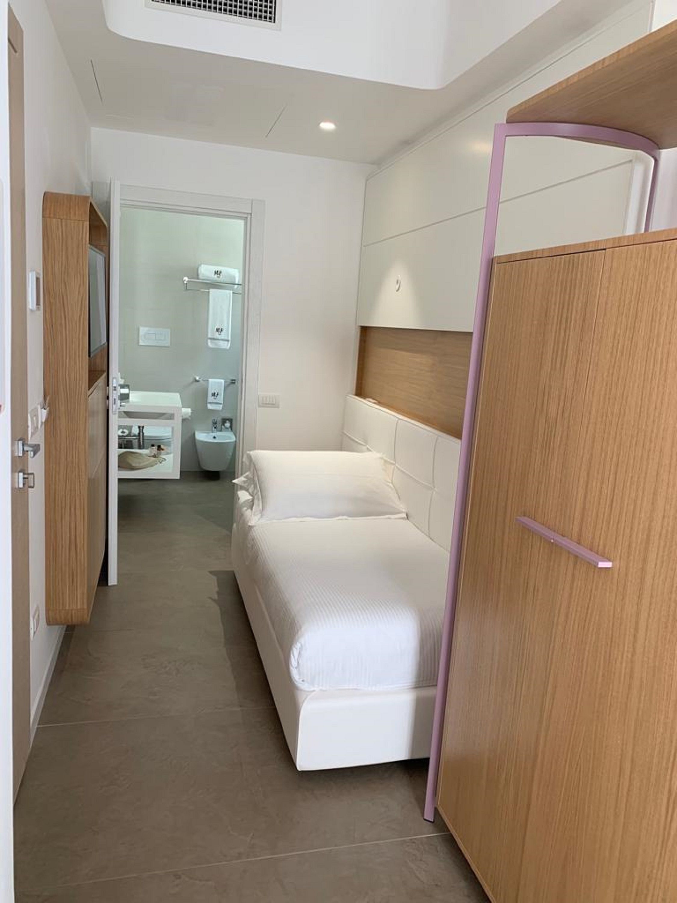 A compact hotel room with a bed, a wooden wardrobe, and a bathroom visible through an open door.