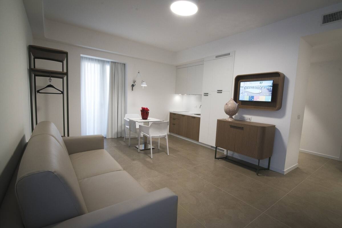Una sala de estar moderna con un sofá, una mesa de comedor con sillas, una zona de cocina con armarios y un televisor montado en la pared.