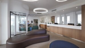 A modern reception area with a curved bench, a reception desk, and a glass door leading to a waiting area. - M&F Hotel (Gallipoli)