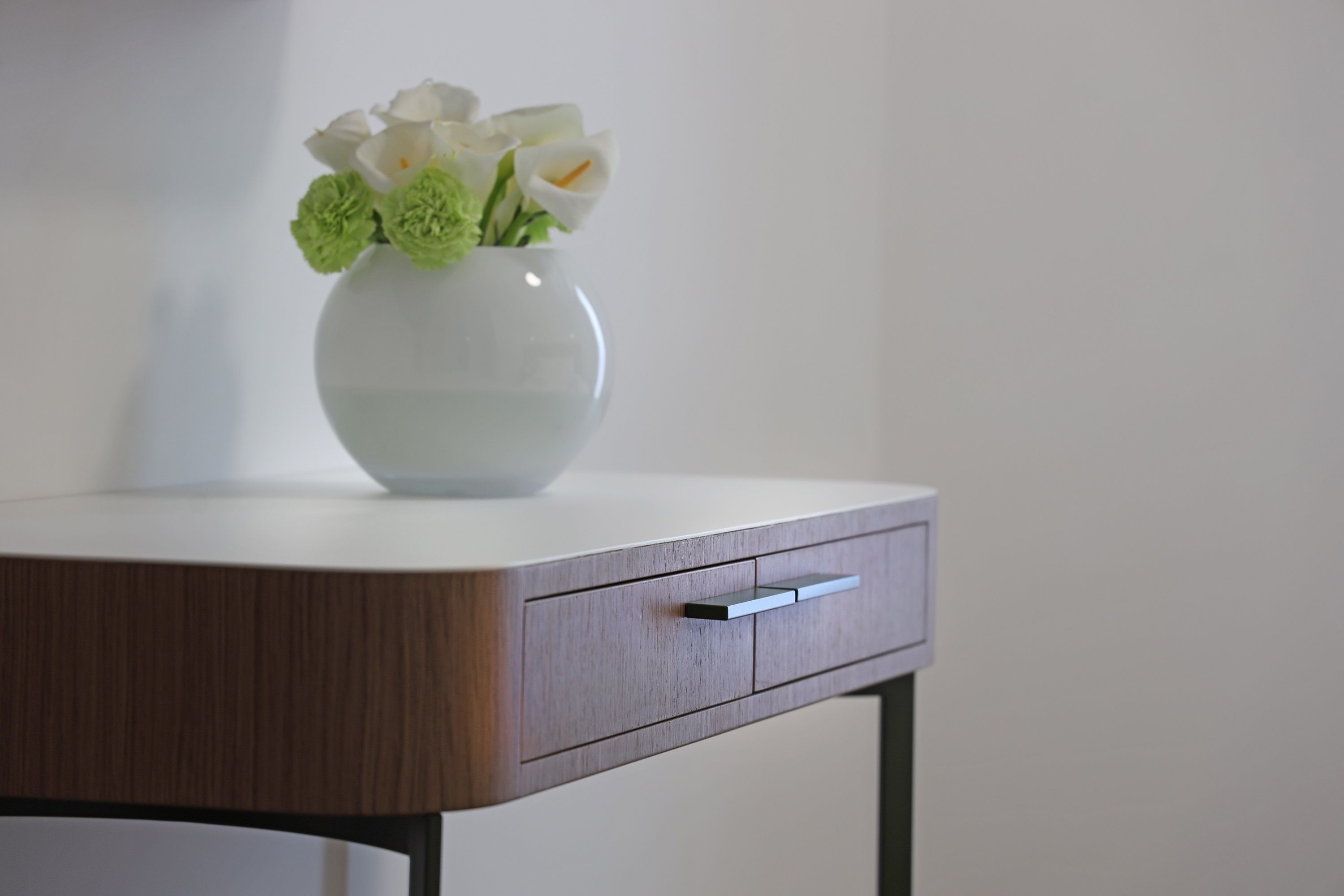 A white vase with flowers on a wooden table against a plain wall.