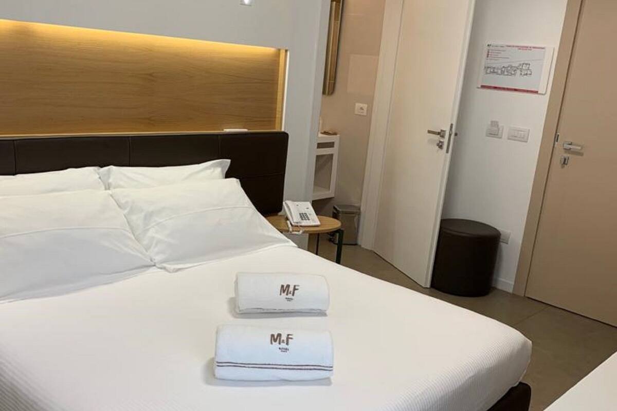 Habitación de hotel con una cama grande, dos toallas blancas con el logotipo 'M.F' y una mesita de noche con un teléfono, además de una puerta con un cartel.