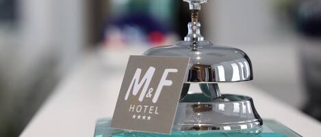 A hotel reception area with a clear glass counter and a silver bell. A sign with "M&F HOTEL" is visible.