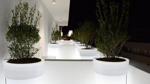 A modern outdoor corridor with white planters containing green plants, leading to a seating area. - M&F Hotel (Gallipoli)