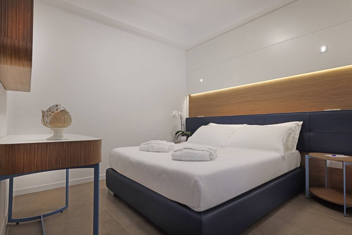 Una habitación de hotel moderna con una cama grande, una mesita de noche, una pequeña mesita auxiliar y un jarrón decorativo.