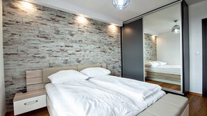 A modern bedroom with a wooden wall, a bed with white bedding, a bedside table, and a large mirror. - Grand Apartments - Bastion Wałowa (Gdansk)