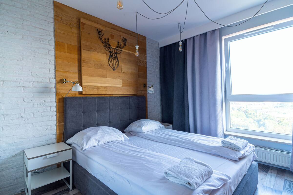 Une chambre moderne avec une tête de lit en bois, deux lits, une table de chevet et une fenêtre avec des rideaux.