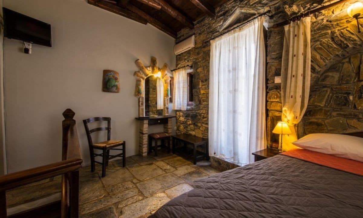 Una habitación rústica con paredes de piedra, una cama con colcha, un escritorio con silla, un espejo y un televisor montado en la pared.