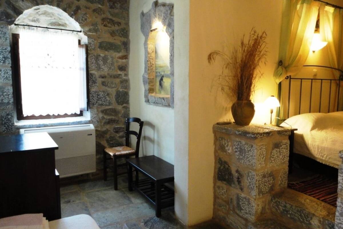 A rustic room with a stone wall, a bed with a metal frame, a bench, a table, and a window with a curtain.