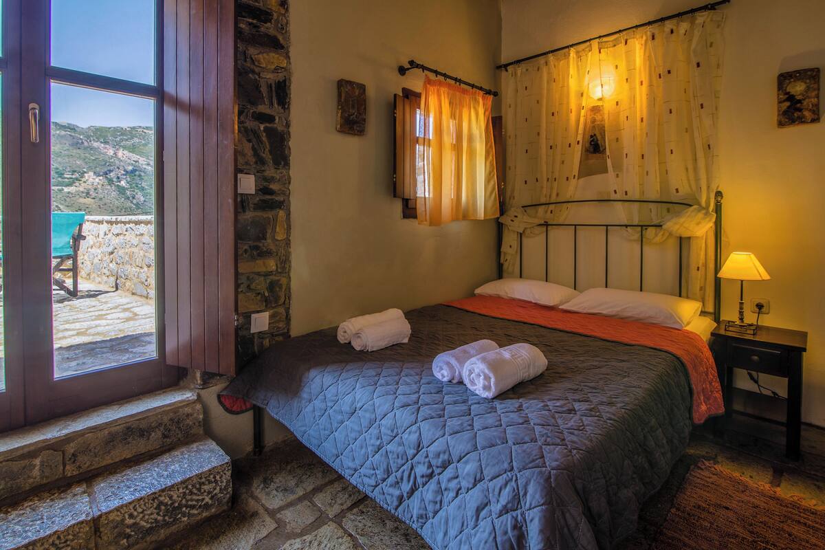 A bedroom with a stone wall, a bed with a quilted blanket, a nightstand with a lamp, and a window with curtains.