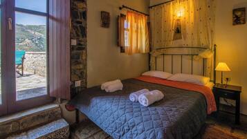 Un dormitorio con pared de piedra, una cama con colcha acolchada, una mesita de noche con lámpara y una ventana con cortinas.