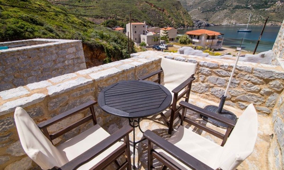A stone balcony with a round table and four chairs, overlooking a coastal village with buildings and a boat in the background.
