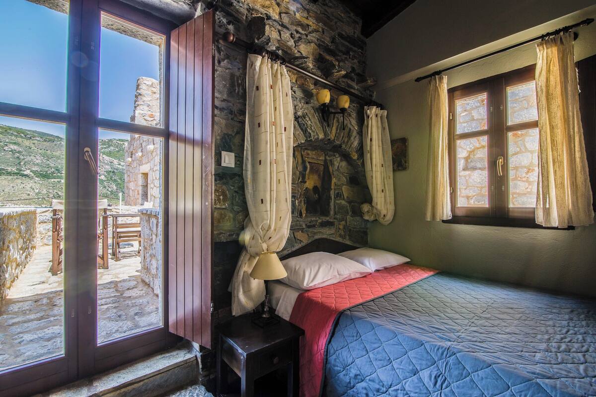 A bedroom with a stone wall, a bed with a red and blue quilt, a nightstand with a lamp, and a view of a stone building outside.